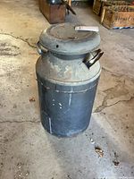 Full view of old metal milk can with lid on, showing wear and rust on surface.