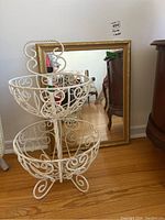 White two-tier metal fruit basket with ornate scroll and bead design in front of a gold-framed mirror.