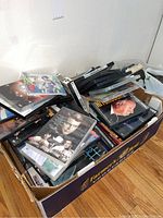 Wide shot of a cardboard box filled with various DVDs in plastic cases and jewel cases. Titles visible include classic films such as Rome Adventure.