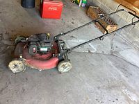 Full view of Canadiana red gas powered lawn mower with black handle, dirty wheels and some grass debris on mower deck.