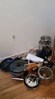 Overall view of aluminum pots, frying pans, crockpot, rolling pins, and utensils stacked on floor