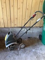 Photo showing the full side profile of the electric roto tiller with handle, small wheels, and plastic housing in a garage setting.