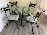 Full view of round glass top table with four matching black wrought iron chairs with curved backs and light gray tufted cushions.