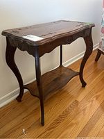 Side view of wooden side table showing curved legs, carved edges, and lower shelf on hardwood floor.