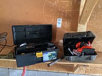 Wide shot of the black plastic toolbox with assorted tools and a case containing the corded drill on a wooden shelf.