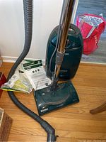 Full view of the Kenmore Elegance green canister vacuum with metal wand attached, hose extending on the floor, and multiple vacuum bags in packaging next to it.
