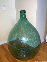 Photo showing front and upper portion of large green glass vase against a beige wall and wood floor.