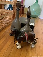 Photo showing two tall glass vases with stoppers and ceramic horse figurine on wooden floor, next to wooden flatware canteen box and a large green glass bottle in background.