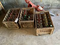 Three vintage wooden Pepsi crates on concrete floor filled with various glass Pepsi bottles. Background shows rough concrete wall and some equipment.