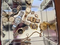 Metal tray holding a collection of men's wristwatches and one pocket watch with chain, showing layout of watches and their different dial colors and bands.