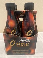Front and top view of four sealed Coca-Cola Blak glass bottles in original cardboard 4-pack carton with Coca-Cola and Blak branding.