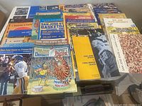 Photo showing a collection of 39 San Jose State University sports programs stacked and spread on a table, featuring covers with various opponents and colorful illustrations and photographs from the 1970s and 1980s.
