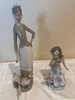 Two Lladro porcelain figurines displayed on a white surface against a wall. The taller one features a woman standing beside two geese; smaller one is a girl kneeling with flowers. Visible damaged geese and missing finger.