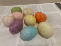 Nine polished stone eggs in assorted colors arranged on a light-colored cloth, showing a variety of smooth, shiny, and colorful eggs.