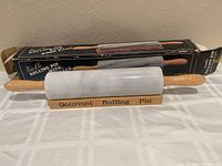 White marble rolling pin with wooden handles resting on curved wooden stand inscribed 'Gourmet Rolling Pin' with the original box placed behind it.