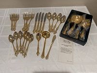 Flatware pieces including forks, knives, spoons laid out on white cloth with serving set in black box and care instructions visible