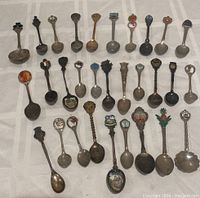 Photo showing all 30 souvenir spoons laid out in rows on a white cloth background, displaying a variety of designs and sizes.