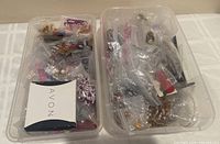 Two clear plastic bins filled with assorted costume jewelry and beads, many pieces in clear plastic bags and one white and navy Avon box visible.