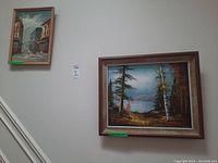 Two framed paintings on canvas hung on staircase wall, one city street scene approx 15x19 inches, one forest and lake landscape approx 20x24 inches