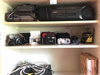 Shelf with multiple cameras including vintage Konica, Kodak, digital camera, Sony CD player, and Sanyo boom box stereo. Also shows camera cases and cables.