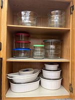Overall view of CorningWare ceramic bakeware and Anchor Hocking glass bowls with lids stored on kitchen cabinet shelves.