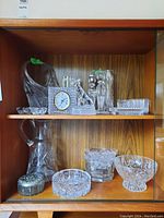 Two wooden shelves with various crystal and glass items including a Mikasa clock, candle holders, bowls, vase, and glass jug.