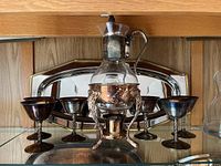 Silver decanter set and six silver goblet-style glasses on warming stand with one silver tray visible in the background.