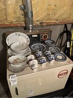 Collection of teacups and plates arranged on a white surface under dim light. Various dishware patterns visible including blue Willow and floral designs.