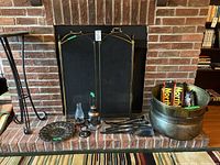 Photo showing black metal fireplace screen with brass handles, fireplace tools including poker, tongs, broom, shovel, metal bucket, and two packs of Northland firelogs, all arranged on a brick fireplace hearth.