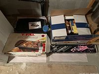 Overview of boxed T-Fal smoke free barbecue, Rival roaster oven, Cuisinart grill and roasting tools in basement setting