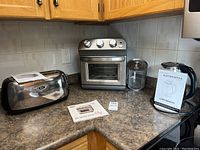 Photo of all three kitchen appliances on a countertop: Oster toaster, Dash air fryer oven with basket, and HadinEEon electric kettle with manual.