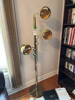 Full view of floor lamp showing triple adjustable shades on brass-finish column and round base; positioned next to bookshelf and window