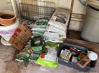 Photo showing bags of soil, grass seed, gardening tools in basket, and planters stacked in garage area.