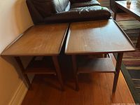 Two square-top teak wood side tables with lower shelf placed side by side next to leather sofa