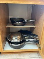 Photo showing stacked pans inside cabinet shelf, includes various sizes of non-stick pans, stainless steel pans, possibly egg poacher and other cookware.