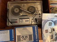 Telefunken reel-to-reel tape recorder with clear dust cover, microphone, and manuals on a wooden surface.