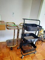 Photo showing both the gold-tone metal serving cart with two glass shelves and the black metal organizer cart with three mesh shelves on caster wheels.