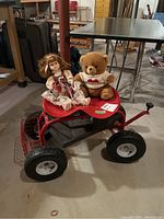 Photo showing red outdoor garden cart with black tires holding a stuffed bear and a porcelain doll sitting on top.