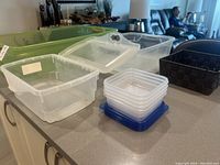 Seven plastic storage containers of various sizes and styles displayed on a countertop including a large green rectangular bucket, two medium translucent buckets with lids, four small square food containers with blue lids, and a small dark brown woven basket.
