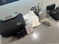 The entire contents of the lot displayed with items spaced out on a grey countertop, showing the digital camera, charger, battery, cables, manual, camera case and pouch.