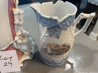 Close-up photo of blue and white porcelain jug with scalloped edge and a painted sailboat scene, showing chips on the top edge