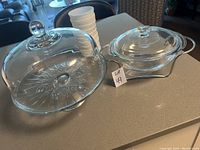 Glass casserole dish with handles and lid placed next to cake dish, along with stacked white cups in background