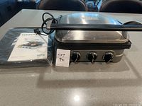 Cuisinart Griddler main unit next to the original manual and spatula on a countertop, front view showing all control knobs.