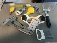 Full set of kitchen utensils including yellow lemon squeezer, herb scissors, salad tongs, lime zester, meat pounder, apple corer displayed on grey countertop