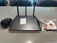 Top view of NetGear Model R7000 router with three antennas standing, power adapter, Ethernet cable, and user manual arranged on a table.