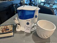 Photo showing Waterpik water flosser with attached handheld flosser, ceramic plant pot with swirl pattern, and a closed box of coasters with teddy bear design, all on a countertop.