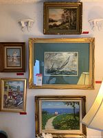 Five framed artworks on a wall including the large sailboat painting at center, top landscape painting, coastal village painting bottom left, figures on beach top left, and pathway with flowers bottom center.