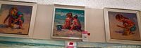 Three framed paintings hanging on wall, showing children playing at the beach in the sand.
