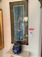 Framed embroidered tapestry on wall with blue ceramic pot with lid and small white wall shelf under it