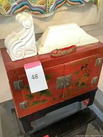 Asian-style cabinet with floral and bird designs on front, metal hardware, standing on black legs with small red and green decorative slipper and two white carved shelves on top.